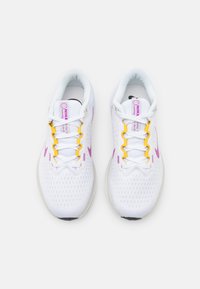 Zapatillas de correr blancas con parte superior de malla transpirable, acentos morados, cordones amarillos y una suela acolchada. Presenta un logo en la lengüeta.