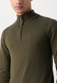 Haut en olive, à manches longues, en pull avec un col zippé moitié. Fabriqué en tissu doux et texturé avec des détails de couture subtils.