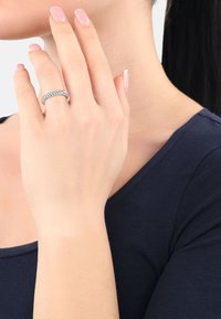 Anillo trenzado de plata en una mano con uñas de color rosa claro, vistiendo una blusa verde marino oscuro. Textura de piel suave y acabado natural de las uñas visibles.
