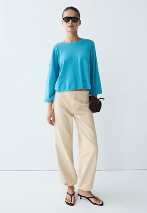 Donna con occhiali da sole neri, top turchese ampio, pantaloni beige a vita alta, sandali neri con tacco, che tiene una piccola borsa marrone.