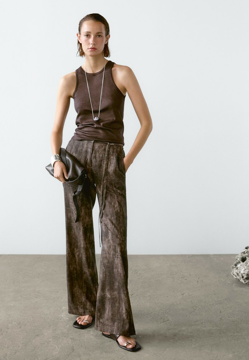Una giovane donna si erge con sicurezza, indossando un top senza maniche scuro e pantaloni larghi e strutturati, tenendo una pochette nera contro uno sfondo minimalista.