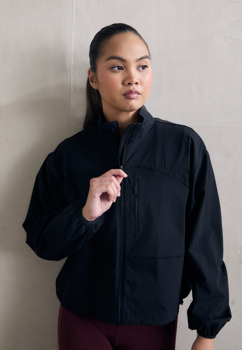 Schwarze Zip-Jacke mit hohem Kragen, elastischen Bündchen und einer vorderen Taschennaht. Hergestellt aus leichtem, strukturiertem Stoff.