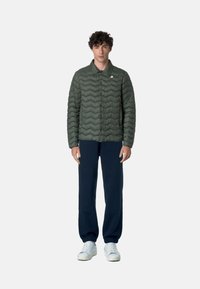 Giacca puffer verde oliva con motivo a onde, design con colletto e chiusura frontale a bottoni a pressione. Abbinata a pantaloni della tuta blu navy e sneakers bianche.