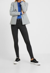 Mujer vestida con jeans skinny negros, zapatillas negras, camiseta azul y chaqueta a rayas gris, de pie con los brazos cruzados.