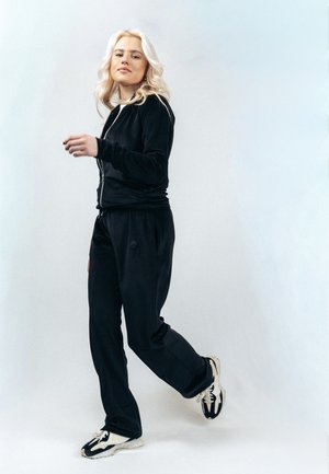 Blondynka w czarnym dresie i trampkach idzie z pewnym siebie wyrazem twarzy na jednolitym jasnym tle.