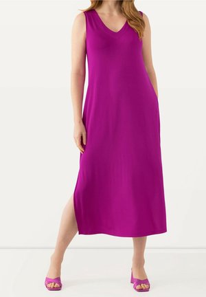 Kobieta ubrana w bezrękawnikową, fuksjową sukienkę midi z dekoltem w serek i rozcięciem z boku, wraz z pasującymi sandałami na obcasie typu blok, stojąca na białej podłodze.