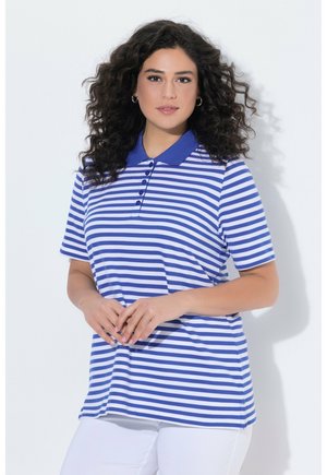 Femme aux cheveux bouclés portant des boucles d'oreilles anneaux et un polo à manches courtes rayé bleu et blanc, posant les mains jointes.