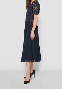 Mörkblå klänning med spetsöverdel och korta ärmar, flytande kjol med lätt genomskinlighet, kombinerad med svarta högklackade skor.