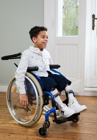 Barn i en blå rullstol med en silverram, klädd i en vit, mönstrad skjorta och mörkblå shorts, vita sneakers och knähöga strumpor.