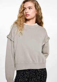 Svetlosivi dukserica s opuštenim krojem, rebrastim izrezom i zavrnutim rukavima. Glatka tekstura, slaže se s crnim uzorcima na donjem dijelu.