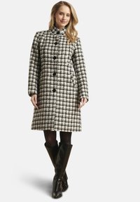 Zwart-witte jas met houndstooth-patroon, voorzien van een hoge kraag, lange mouwen en zes grote knopen aan de voorkant. Draagt daarbij kniehoge zwarte laarzen.