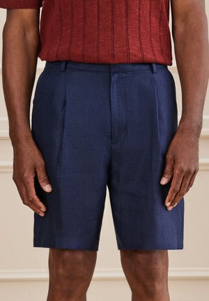 Mann trägt blaue maßgeschneiderte Shorts und ein rotes kurzärmeliges Strickhemd und steht vor einem beigen Hintergrund.
