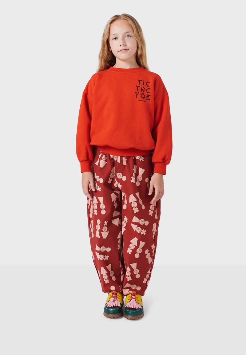 Rode sweatshirt met bedrukte tekst, gecombineerd met losse, gestreepte rode broek met lichtroze vormen. Multicolour schoenen met franjes.