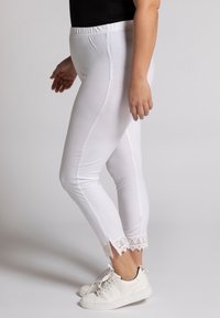 Woman wearing white fitted leggings with lace trim and side slits at ankles, paired with white sneakers and a black top.