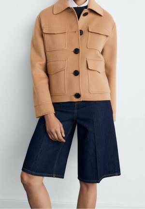 Personne portant une veste boutonnée couleur chameau avec quatre poches frontales et un short en jean large bleu foncé jusqu'aux genoux.