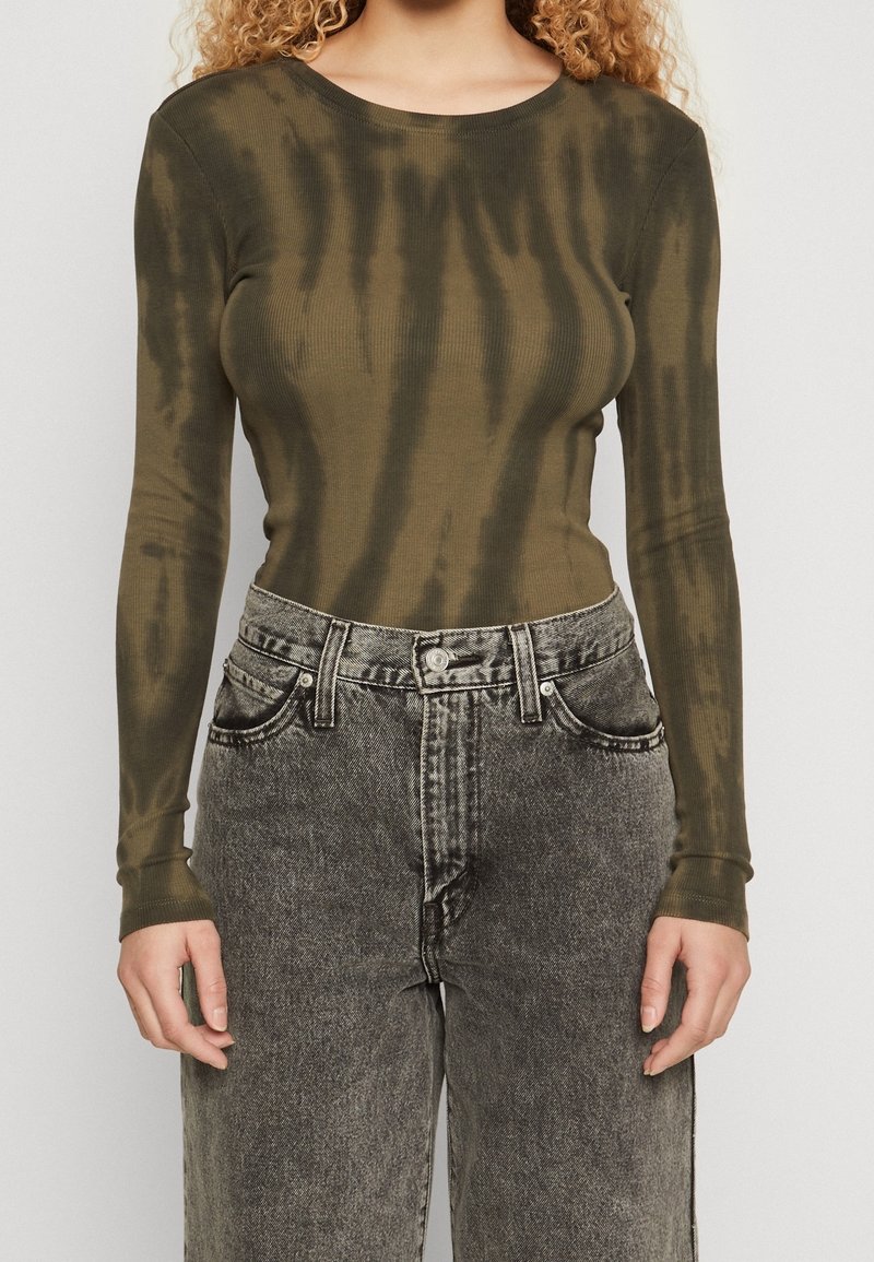 Femme portant un haut tie-dye olive vert à manches longues rentré dans un jean en denim noir délavé taille haute, se tenant contre un fond uni.
