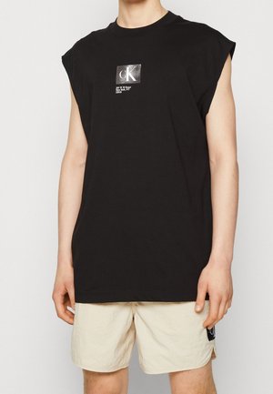 Person in einem schwarzen ärmellosen CK-T-Shirt mit weißem Text und beigen Shorts vor einem einfarbigen Hintergrund.