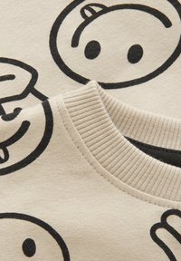 Beige sweatshirtstof med sort tegneserie-smiley-ansigt og håndgrafik. Ribbet kravedetalje. Blød og struktureret udseende.