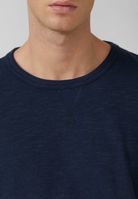 Close-up van de nek en onderkant van het gezicht van een persoon die een donkerblauw, gestructureerd shirt met ronde hals draagt, met een gestikte V-vormige detail onder de kraag.