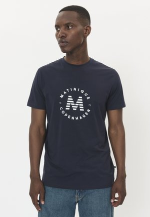 Marineblaues Baumwoll-T-Shirt mit einem kreisförmigen weißen Logo, das "MATINIQUE COPENHAGEN" und einen gestreiften Großbuchstaben "M" zeigt. Mit kurzen Ärmeln und Rundhalsausschnitt.