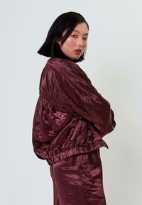 Burgundi satino bomberio striukė su elastingomis rankovėmis ir apvadu. Yra sukaupto audinio detalės petyse ir laisvas kirpimas. Lygus paviršius.