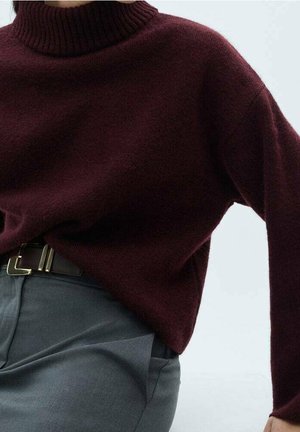 Person iført en mørk burgundy rullekravesweater stukket ned i grå skræddersyede bukser med sort bælte mod en ensfarvet baggrund.