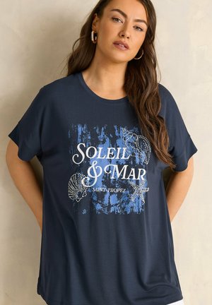 Pitkät ruskeat hiukset omaava nainen, jolla on päällään laivastonsininen T-paita, jossa lukee "SOLEIL & MAR SAINT TROPEZ" ja joka on koristeltu simpukka- ja meritähdellä.