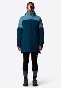 Teal og mørkeblå vandtæt jakke med hætte, to frontlommer, båret over sorte leggings og mørke sko, ledsaget af reflekterende solbriller.