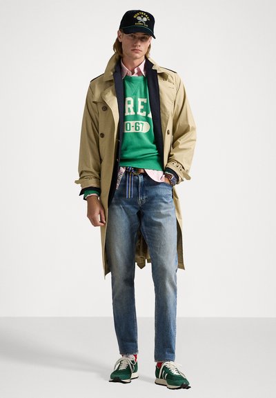 Un trench coat beige sopra un felpa verde, jeans blu e sneakers verdi. L'outfit include un cappellino con logo vintage e una cintura multistrato.