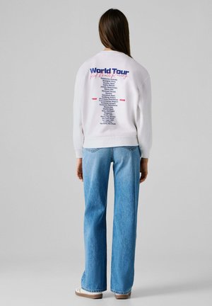 Mujer con sudadera blanca y vaqueros azules, la parte trasera de la sudadera muestra el texto "Gira Mundial" con una lista de ciudades globales y años.