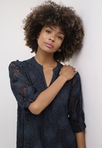 Tengerés kék blúz csipkés díszítéssel, puffos ujjakkal, V-kivágással és virágmintákkal. Sima textúra bonyolult hímzéssel díszítve.