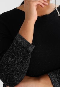 Maglione nero con un polsino testurizzato lucido nero e argento. Presenta un collo a coste e una silhouette aderente.