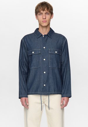Giovane uomo che indossa una camicia di denim scuro abbottonata con due tasche sul petto e pantaloni chiari, in piedi davanti a uno sfondo semplice.