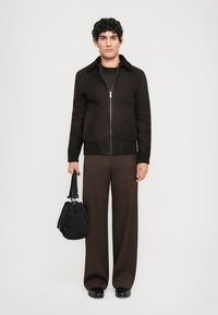 Jachetă crem cu fermoar, cu o suprafață texturată și guler negru, asortată cu pantaloni lărgi, bej. Persoana ține o geantă neagră.