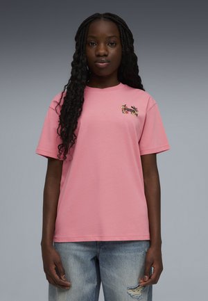 Joven mujer con camiseta rosa de Puma con logo floral en el pecho y jeans azules rasgados, posando frente a un fondo gris.