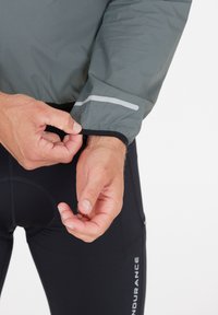 Leichte graue Jacke mit elastischen Bündchen und reflektierendem Streifen. Getragen über schwarzen Sporthosen mit sichtbarem Logo.