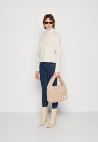 Sac à main en peluche beige avec deux poignées courtes, associé à un pull col roulé crème et un jean skinny bleu. Le mannequin porte des bottines beige clair.