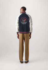 Chaqueta varsity en azul marino y crema con puños y dobladillo acanalados, que presenta un logo circular de "Nueva York" y pantalones marrones, usados con zapatillas casuales.