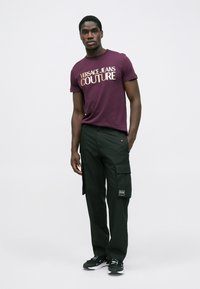 Jauns vīrietis tērpies violetā "Versace Jeans Couture" T-kreklā, melnās kargo biksēs ar logo, un melnās sporta apavos, stāv uz viendabīga fona.