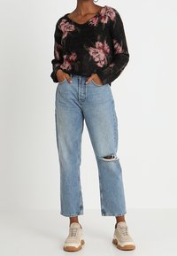 Maglione nero con motivo floreale rosa, scollo a V e vestibilità ampia. Jeans azzurri, a vita alta, con strappo al ginocchio. Sneakers beige con suole bianche.