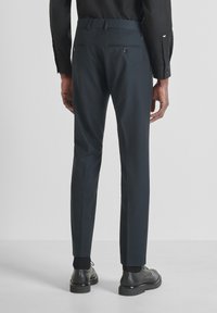 Man in een slim-fit, donkergrijze pantalon, zwart overhemd, zwarte sokken en zwarte leren nette schoenen, staand op een lichtgrijze vloer.