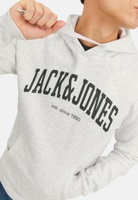Lysgrå hoodie med en kengurulomme, der har fed sort tekst "JACK&JONES" og "intl. siden 1990" trykt på brystet.