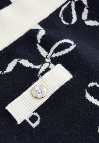 Un tessuto lavorato a maglia navy e bianco con un motivo a nastro bianco e una tracolla rigata bianca fissata da un bottone con bordo dorato e testo.