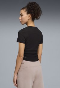 Νεαρή γυναίκα με σγουρά μαλλιά σε αλογοουρά φοράει μαύρο μπλουζάκι με κοντά μανίκια και μπεζ παντελόνι, στέκεται μπροστά σε απλό γκρι φόντο.