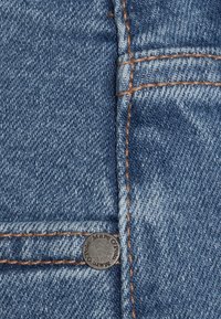 Gros plan sur un jean en denim bleu avec des coutures orange et un rivet métallique sur la couture d'une poche.