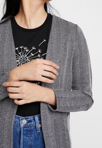 Černobílé herringbone kardigan s otevřeným předním dílem a tříčtvrtečními rukávy, nošený přes černé tričko s potiskem a džíny s vysokým pasem.