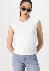 Top sem mangas branco canelado com decote redondo, combinado com jeans de denim azul claro. O tecido tem um acabamento texturizado.