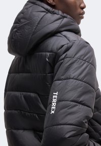 Personne portant une veste matelassée noire à capuche avec « TERREX » brodé en blanc sur la manche droite, vue de profil et de dos.