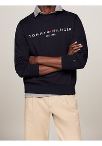 Uomo che indossa una felpa navy di Tommy Hilfiger sopra una camicia grigia con pantaloni beige, braccia incrociate in vita.
