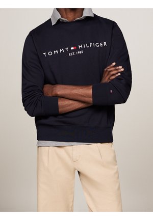 Hombre vestido con un sudadera navy de Tommy Hilfiger sobre una camisa de color gris con pantalones beige, con los brazos cruzados a la altura de la cintura.